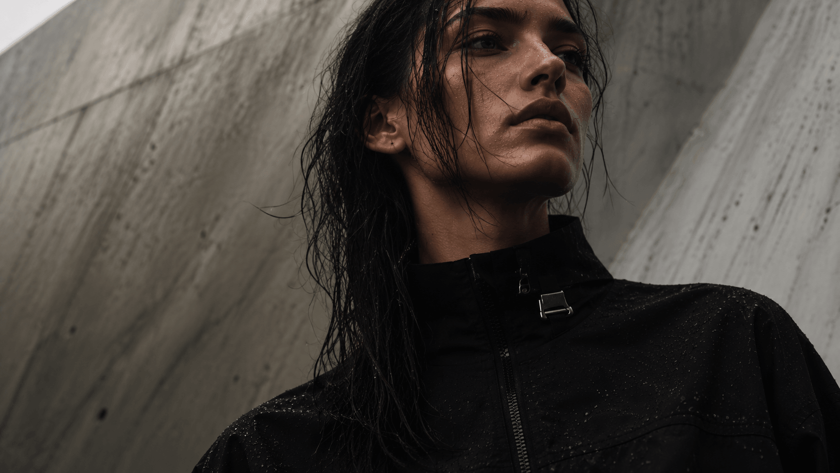 A model with wet hair in a black shell jacket against a concrete wall